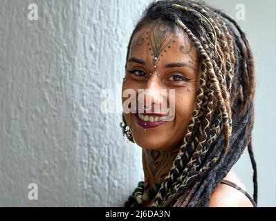 Une jeune femme mexicaine alternative attrayante avec de longues tresses rasta, un piercing facial d'art corporel et des sourires de tatouage positivement optimiste au spectateur. Banque D'Images