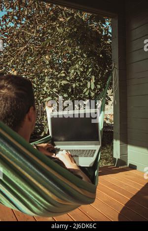 Un homme indépendant se trouve dans un hamac et travaille près de la maison de campagne par une journée ensoleillée. Travail à distance Banque D'Images