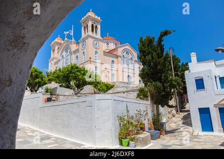 Tinos, Grèce - 1 juillet 2021 : église traditionnelle avec maçonnerie rose, grande horloge de la tour et un drapeau de la Grèce agitant dans le vent. Vue à travers une arcade, en montée Banque D'Images