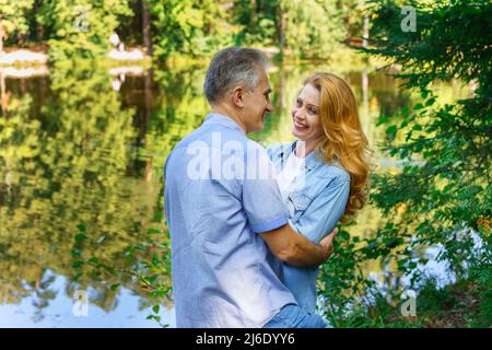 Un couple heureux et mûr qui s'embrasse dans une tournière épaisse près du lac sourient avec bonheur lors d'une journée ensoleillée d'été. L'homme et la femme caucasiens passent du temps libre ensemble Banque D'Images