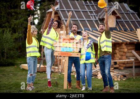 Un groupe de jeunes hommes et femmes de construction joyeux sont excités tout en posant pour une photo à un chantier de construction de chalet sur une belle journée. Constr Banque D'Images