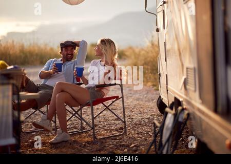 Couple au coucher du soleil en applaudirant avec des boissons. Assis devant le camping-car. Amusant, ensemble, concept de nature. Banque D'Images