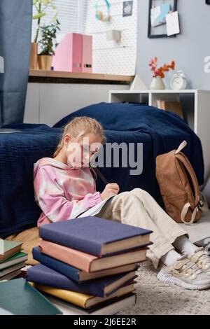Écolière concentrée à capuche assise sur le sol derrière une pile de livres et se préparant à l'examen Banque D'Images