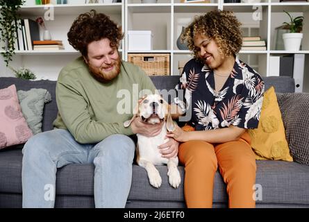 Un jeune couple heureux assis sur un canapé avec son chien, ils caressent leur animal de compagnie et parlent avec lui Banque D'Images