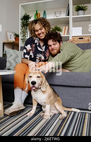 Heureux multiethniccouple passer du temps libre sur le canapé avec leur chien assis sur le sol et profiter de leur affection Banque D'Images