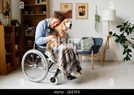 Un jeune barbu s'embrasse avec son joli chien tout en étant assis en fauteuil roulant dans la salle de séjour Banque D'Images
