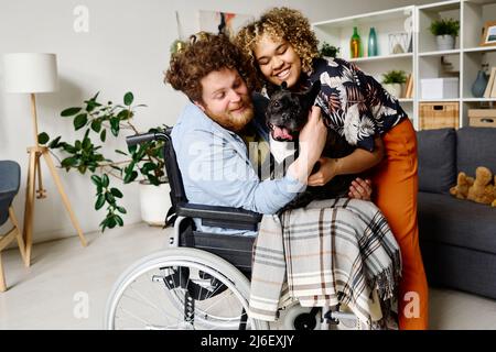 Jeune barbu en fauteuil roulant passant la réhabilitation à la maison avec sa femme et son chien charmant Banque D'Images