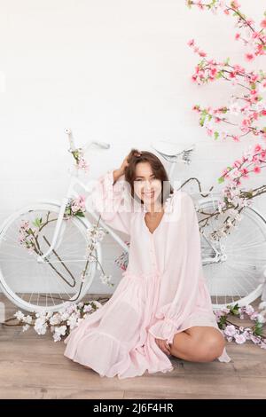 Portrait d'une jeune femme heureuse avec de longs cheveux portant une robe rose, assise près d'un vélo blanc parmi des fleurs artificielles. Banque D'Images