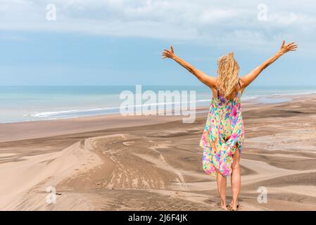 Photo avec espace de copie de l'arrière d'une femme blonde levant les bras sur le dessus d'une dune face à l'océan Banque D'Images