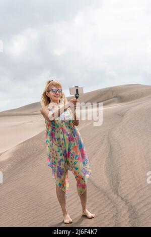 Photo verticale d'une femme avec des lunettes de soleil à l'aide d'un trépied portable pour prendre un selfie dans une dune Banque D'Images