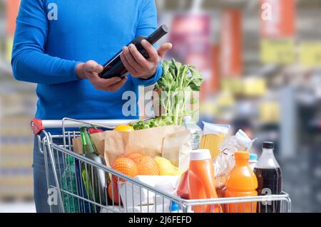 Homme faisant des courses d'épicerie au supermarché, il achète une bouteille de vin rouge, panier complet au premier plan Banque D'Images