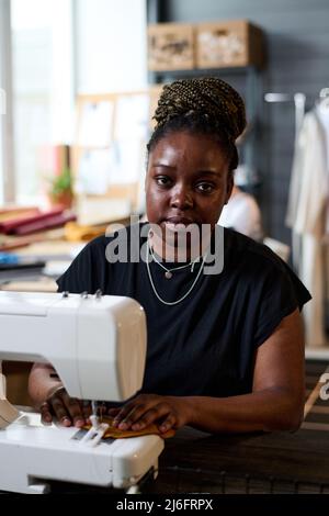 Jeune femme noire regardant l'appareil photo tout en étant assise sur le lieu de travail et en utilisant la machine à coudre électrique pour créer de nouveaux vêtements Banque D'Images