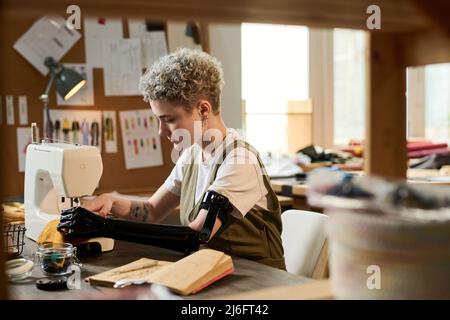 Jeune femme sérieuse tailleur avec bras partiel se concentrant sur le travail de couture tout en étant assis près d'une table devant la machine électrique Banque D'Images