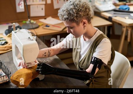 Jeune couturière sérieuse avec prothèse de bras noir travaillant par machine à coudre électrique tout en créant des vêtements pour la nouvelle collection de mode Banque D'Images