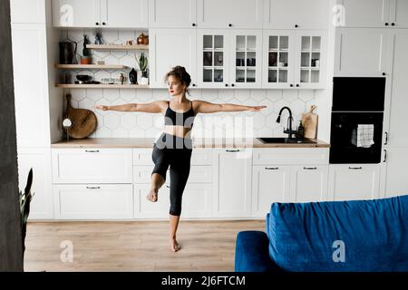 Entraîneur sportif de forme physique de fille montre des leçons en ligne. Jeune fille en forme de curly saine faisant du yoga à la maison tout en se tenant dans une posture d'arbre, concept de mode de vie sain Banque D'Images