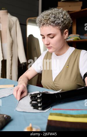 Jeune couturière sérieuse circling papier modèle de nouveaux vêtements par son lieu de travail tout en travaillant sur ordre de client ou collection de mode Banque D'Images