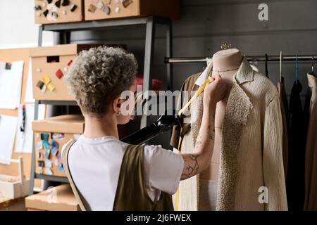 Vue arrière des jeunes marins prenant des mesures de manteau beige sur mannequin tout en travaillant sur un nouvel ordre de client dans l'atelier Banque D'Images