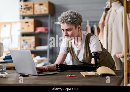 Jeune designer de mode sérieux regardant à travers la nouvelle tenue tendance sur l'écran d'ordinateur portable tout en travaillant sur la collection saisonnière dans l'atelier Banque D'Images