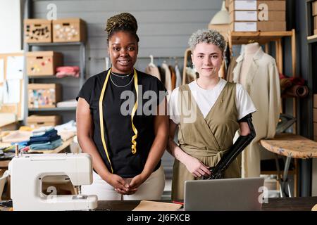 Deux jeunes créateurs de mode interculturels heureux debout sur le lieu de travail équipé d'une machine à coudre électrique dans un atelier spacieux Banque D'Images