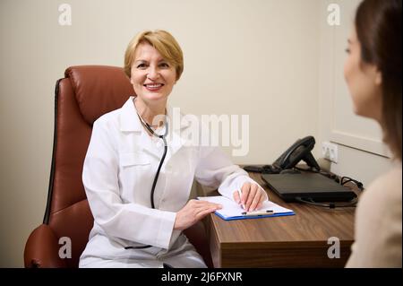 Confiante mature femme généraliste, médecin, médecin remplissant un formulaire médical tout en consultant un jeune patient en clinique et cutely smili Banque D'Images