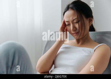 Migraine. Vue latérale de la souffrance malheureuse de mal de tête bronzé belle jeune femme asiatique touchant le front à la maison intérieur salon. Blessures Banque D'Images