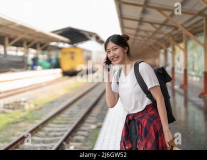 Belle jeune femme asiatique avec un sac à dos utilise le téléphone tout en se tenant près du train de chemin de fer sur la plate-forme. Concept d'été de voyage bon marché Banque D'Images