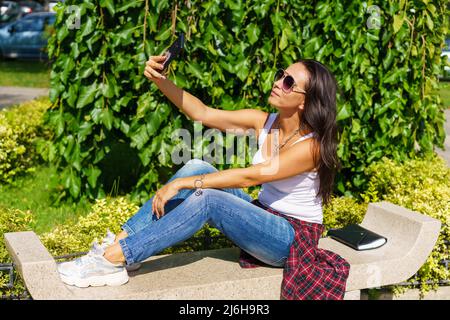 Une jeune femme gaie prend le selfie des mains avec le téléphone tout en étant assise sur le banc de parc ou sur le fond de jardin. Portrait jeune femme attrayante faisant selfie photo sur smartphone le jour de l'été en lunettes de soleil Banque D'Images