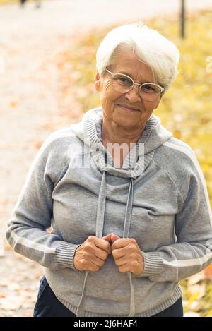 Femme âgée de race blanche debout dans un parc, regardant la caméra, souriant et touchant son sweat à capuche gris. Prise de vue verticale en extérieur. Photo de haute qualité Banque D'Images