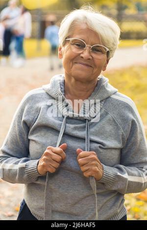 Bonne vieille femme caucasienne avec des lunettes en gris portrait à capuche dans le parc, jour d'automne. Tir vertical. Photo de haute qualité Banque D'Images