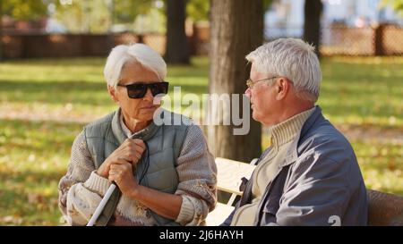 Grands-parents européens retraités à poil gris assis sur un banc dans un parc en automne. Invalide aveugle caucasienne femme senior avec grue blanche et lunettes de soleil parle avec son partenaire. Photo de haute qualité Banque D'Images