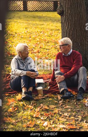 Tir vertical. Couple caucasien âgé assis sur une couverture au parc, buvant du café, parlant, partageant des souvenirs précieux. Concept pique-nique. Photo de haute qualité Banque D'Images
