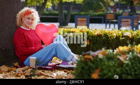 Belle fille blanche blonde aux cheveux bouclés tenant un ballon rouge en forme de cœur et regardant la caméra tout en étant assise sur les feuilles d'automne sous un arbre. Photo de haute qualité Banque D'Images