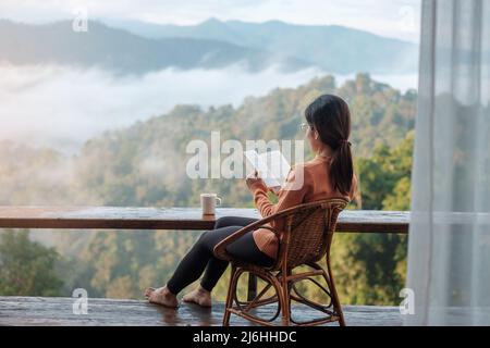 jeune femme lisant le livre près de la fenêtre et regardant la vue sur la montagne à la campagne homestay dans le lever du soleil matin. SoloTravel, voyage, voyage et détente Banque D'Images