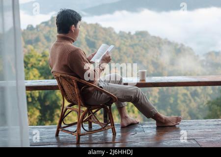 jeune homme lisant le livre près de la fenêtre et regardant la vue sur la montagne à la campagne homestay dans le lever du soleil matin. SoloTravel, voyage, voyage et détente co Banque D'Images