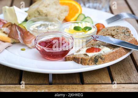 Petit-déjeuner avec pain, beurre et confiture, aussi diverses viandes froides et fruits sur une assiette blanche sur une table extérieure en bois, sélection de focus, profondeur étroite de fi Banque D'Images