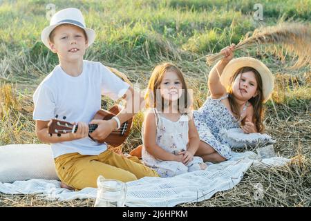 Portrait d'un garçon joyeux et de deux filles assis sur une couverture dans le champ, ayant pique-nique.Un moment de détente.Jouer de la guitare, ukulele, parler, plaisanter.Lumière ensoleillée Banque D'Images