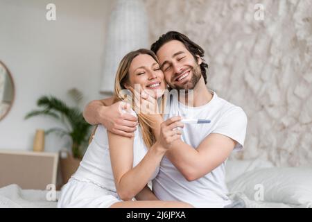 Un jeune couple heureux qui célèbre le résultat positif du test de grossesse, qui s'embrasse et se câlin sur le lit à la maison Banque D'Images