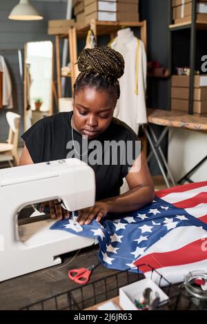 Jeune femme noire sérieuse en t-shirt bleu couture drapeau américain tout en étant assis devant la machine électrique par une grande table dans l'atelier Banque D'Images