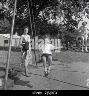 1950s, historique, un jeune garçon et une fille sur le terrain de jeu balançoires, avec leur mère dans une robe à motif fleuri debout en regardant, Angleterre, Royaume-Uni. Les deux enfants jouent sur des balançoires traditionnelles avec des sièges en bois suspendus sur des chaînes métalliques attachées à un cadre en métal sur une base en béton. Banque D'Images