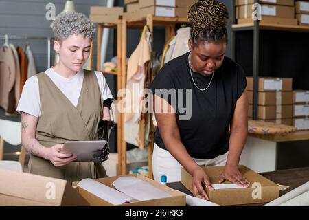 Une employée d'entrepôt qui vérifie les commandes en ligne avec une tablette tout en aidant sa collègue afro-américaine à apposer un autocollant avec adresse sur la boîte Banque D'Images