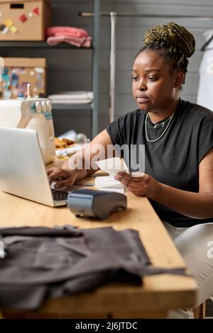 Jeune femme sérieuse d'entrepôt vérifiant les retraits de paiement pour les commandes en ligne tout en étant assis sur le lieu de travail devant l'ordinateur portable Banque D'Images