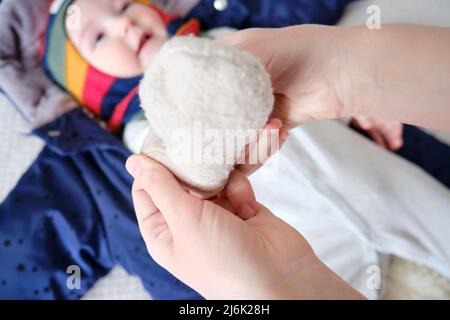 Une mère femme met des moufles chaudes pour un bébé garçon. Maman habille un enfant heureux dans des vêtements d'hiver sur le lit. Banque D'Images