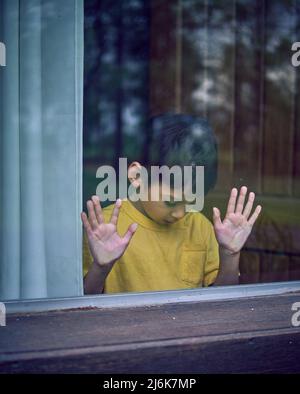 Seul enfant latino pendu ses mains derrière le verre de la fenêtre. Petit garçon regardant vers le bas ennuyé et triste à l'intérieur de sa maison. Verticale Banque D'Images