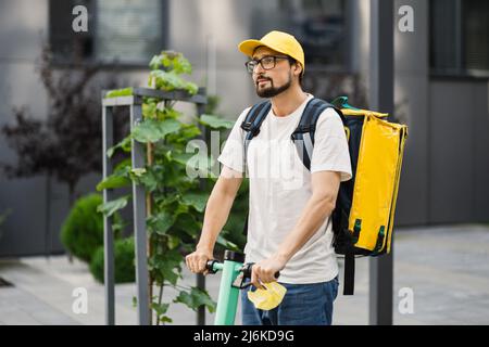 Cavalier barbu homme dans la casquette jaune et sac à dos de boîte thermique livrant le repas aux clients avec le scooter électrique. Concept alimentaire écologique à livraison rapide. Banque D'Images