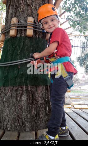 Un petit garçon dans un parc d'attractions dans un harnais de sécurité sur un arbre surmonte un obstacle Banque D'Images