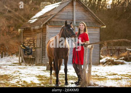 Une belle fille dans une robe rouge marche à travers une vieille ferme en hiver dans les rayons du soleil couchant Banque D'Images