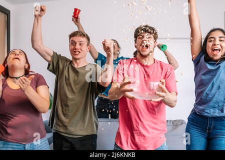 les jeunes crient avec enthousiasme en lançant du pop-corn dans les airs après la victoire de leur équipe, diffusée à la télévision. Banque D'Images