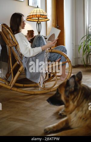 Jeune femme lisant un livre avec chien Berger allemand, assise sur une chaise à bascule. Ambiance chaleureuse, vie lente, personnes avec animaux de compagnie. Maison d'époque Banque D'Images