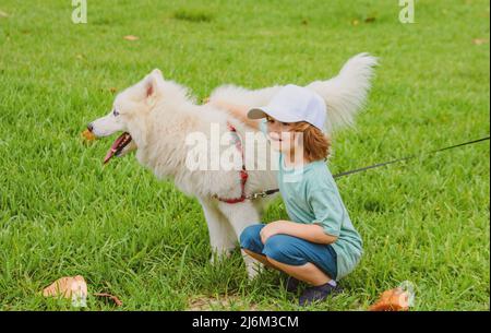 Un petit garçon enchâssant un chien blanc doux et doux qui rit dans le parc ou qui pousse l'herbe sur fond. Banque D'Images