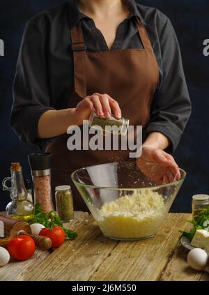 Un chef professionnel prépare des plats au fromage sur une table de cuisine en bois. Ajoute des épices au fromage râpé. Beaucoup d'ingrédients sur la table - légumes Banque D'Images